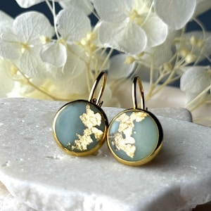 Könnte beinhalten: Ein Paar goldfarbene Ohrringe mit einem runden, hellblauen Mittelteil und Goldblatt-Akzenten. Die Ohrringe haben einen Klappbügelverschluss. Der Hintergrund zeigt weiße Blüten.