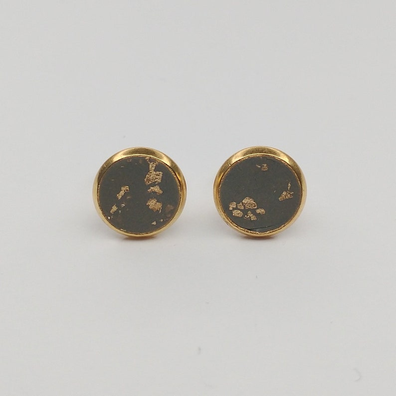 May include: A pair of gold stud earrings with a dark gray background and gold flecks.