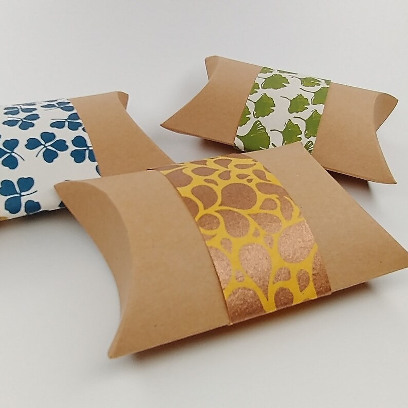 May include: Three pillow boxes made of brown cardboard with a decorative paper band. The boxes have different patterns on the bands: blue clover leaves, green ginkgo leaves, and gold paisley.