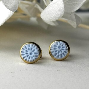 Könnte beinhalten: Ein Paar Ohrstecker mit hellblauem Blumenmuster, gefasst in goldfarbenem Metall. Die Ohrringe haben eine runde Form und ein filigranes, detailliertes Muster. Die Ohrringe werden auf neutralem Hintergrund präsentiert.