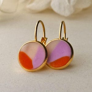 Könnte beinhalten: Ein Paar Ohrringe mit Brisurverschluss und runden Anhängern. Jeder Anhänger zeigt eine glatte, mehrfarbige Einlage mit deutlichen Abschnitten in leuchtendem Orange, zartem Lavendel und Creme, eingefasst in einen polierten, goldfarbenen Metallrahmen.