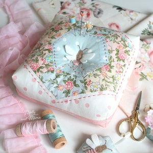 May include: A white pincushion with a hexagonal patchwork design featuring a bee and floral patterns. The pincushion is surrounded by pink lace, spools of thread, and a pair of gold scissors. The text "Molly and Mama" is visible in the lower left corner.