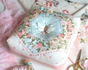 May include: A white pincushion with a hexagonal patchwork design featuring a bee and floral patterns. The pincushion is surrounded by pink lace, spools of thread, and a pair of gold scissors. The text "Molly and Mama" is visible in the lower left corner.