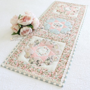 May include: A pink and blue floral table runner with a white lace trim. The runner is made of fabric squares sewn together in a patchwork pattern. The runner is on a white surface.
