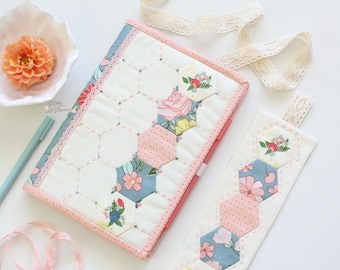 May include: A white fabric journal cover with a pink border and a hexagonal pattern of floral fabric. The cover has a matching bookmark with a pink and blue floral pattern.
