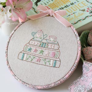 Puede incluir: Un aro de bordado rosa y blanco con un diseño de una pila de libros con tazas de té en la parte superior. Los libros están bordados con hilos de diferentes colores y las tazas de té son rosas con corazones. El aro está sobre una tela rosa y blanca.