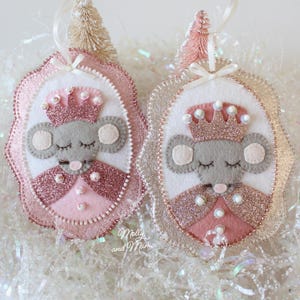 May include: Two felt Christmas ornaments. Each features a grey mouse wearing a pink crown and dress. The ornaments have a glittery pink and silver border, white accents, and a ribbon for hanging. Decorated with pearls and small Christmas trees.