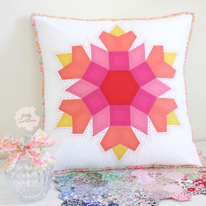 Może przedstawiać: Biała poduszka z geometrycznym wzorem kwiatu w kolorze różowym, pomarańczowym i żółtym. Poduszka ma białe tło i geometryczny wzór kwiatu w kolorze różowym, pomarańczowym i żółtym w środku. Poduszka jest otoczona białą ramką z geometrycznym wzorem w kolorze różowym, pomarańczowym i żółtym.