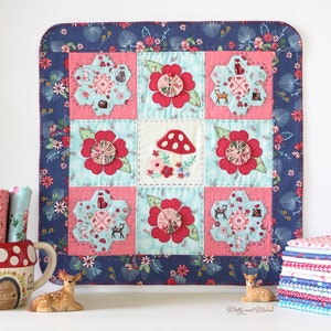 May include: A handmade quilt block with a blue floral border and a central design of red and pink flowers, a mushroom, and a deer. The quilt block is made with a variety of fabrics and is quilted with a simple stitch.