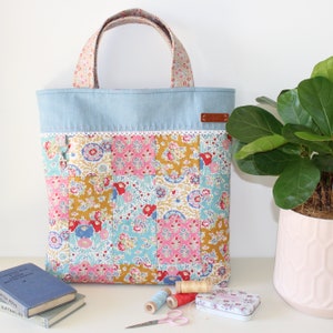 May include: A blue and white patchwork tote bag with a floral print. The bag has a brown leather handle and a white lace trim. The bag is sitting on a table with a plant and some sewing supplies.