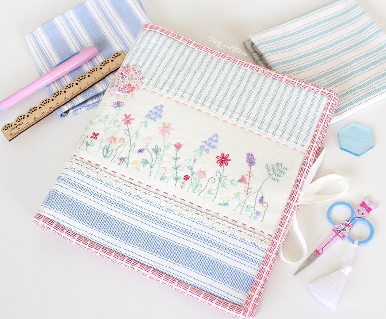 May include: A white fabric storage box with a blue and white striped pattern and a pink border. The box has a floral embroidery design and a white lace trim. The box is closed with a white ribbon.