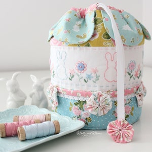 Może przedstawiać: Materiałowy koszyk wielkanocny z kwiatowym i króliczym wzorem. Koszyk ma jasnoniebieską i różową kolorystykę, z białą wstążką i ozdobnym kwiatkiem na dole. Koszyk jest ozdobiony haftowanymi króliczkami i kwiatami.