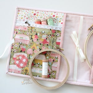 May include: Pink fabric storage case with a zippered pocket and two embroidery hoops. The fabric features a floral pattern with vintage campers. The case has a white ribbon tied in a bow.