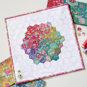 May include: A white fabric square with a pink border, featuring a colourful hexagonal quilt pattern in the centre. The quilt is made of various floral fabrics and is surrounded by a white background with a subtle grid pattern.