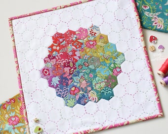 May include: A white fabric square with a pink border, featuring a colorful hexagonal quilt pattern in the center. The quilt is made of various floral fabrics and is surrounded by a white background with a subtle grid pattern.
