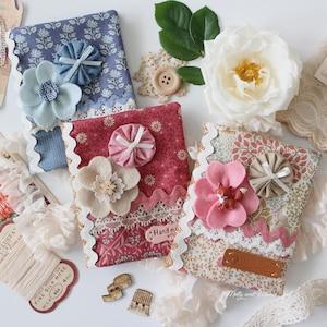 May include: Handmade fabric notebooks with floral and lace details. The notebooks display floral patterns in blue, red, and floral prints. Each notebook is decorated with a felt flower and decorative trim. The image includes sewing notions and a white rose.