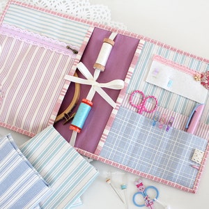 May include: A pink and blue striped fabric organizer with multiple pockets and compartments. The organizer is filled with sewing supplies, including scissors, thread, needles, and a hoop.