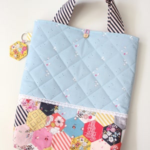 Puede incluir: Una bolsa de mano acolchada de color azul claro con un asa de rayas blancas y negras. La bolsa tiene un diseño de patchwork en la parte inferior con un ribete de encaje blanco. La bolsa tiene un pequeño llavero en forma de hexágono unido a ella.