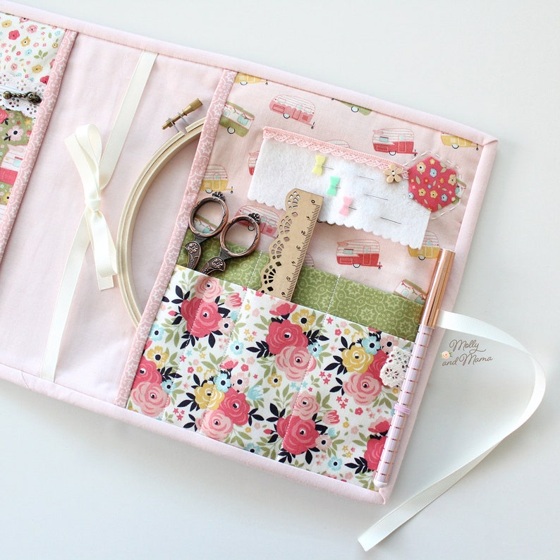 May include: A pink fabric sewing kit with multiple pockets and compartments. The kit includes a wooden embroidery hoop, scissors, a ruler, pins, and a pen. The fabric is patterned with pink and white flowers and has a small label that says "Molly and Mama".