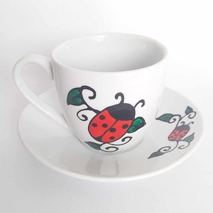 May include: A white teacup and saucer with a hand-painted design of ladybirds and leaves. The ladybirds are red with black spots and the leaves are green.