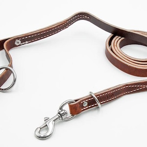 May include: A brown leather dog leash featuring white contrast stitching, a silver-toned metal swivel snap hook, and a circular metal ring near the handle. The leash is coiled at one end and includes metal rivets securing the leather loops.