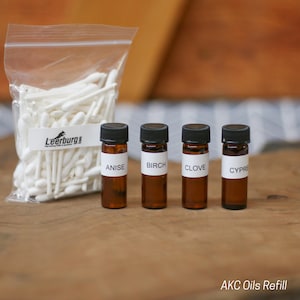 May include: Four small brown glass bottles with white labels. The labels read "Anise", "Birch", "Clove", and "Cypre". A bag of white cotton swabs is in the background. The text "AKC Oils Refill" is at the bottom of the image.