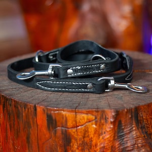 Puede incluir: Juego de correa y collar para perro de cuero negro con herrajes plateados. La correa y el collar están cosidos con hilo blanco y presentan el texto "Loorburg.com". El conjunto se muestra sobre una superficie de madera.
