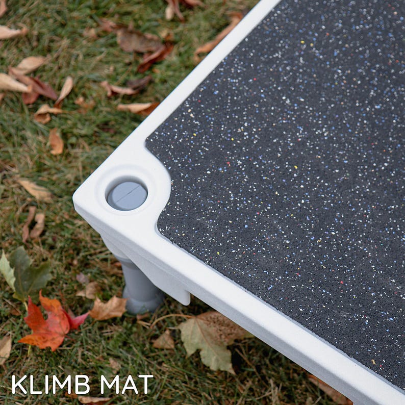 Puede incluir: Escal&oacute;n blanco para mascotas con una superficie texturizada negra y patas de pl&aacute;stico gris. El escal&oacute;n tiene una tapa de pl&aacute;stico gris en la esquina. El texto "KLIMB MAT" es visible en la esquina inferior izquierda de la imagen.