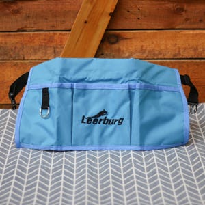 以下が含まれることがあります： 複数のポケットと黒いLeerburgロゴが付いた薄い青色のユーティリティベルト。ベルトには金属製のDリングと調節可能なストラップが付いています。