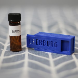 May include: A small brown glass bottle with a white label that says "Birch" next to a blue plastic container with the word "LEERBURG" printed on it.