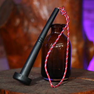 May include: A black fire starter with a textured handle rests next to a brown glass bottle. A red, white, and blue braided cord is looped around the bottle's neck. The fire starter has the text "UNEX 20" on the base.
