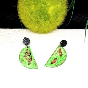Pode incluir: Brincos feitos à mão com um design semicircular verde limão e um pino preto. Os brincos apresentam uma pequena ilustração abstrata em vermelho e castanho. Em um fundo branco com elementos decorativos verdes e amarelos.