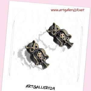 Peut inclure: Une paire de boucles d'oreilles en forme de hibou en bronze. Les hiboux sont tournés vers l'avant, les ailes déployées. Les boucles d'oreilles sont sur un fond blanc avec le texte "www.artgallery29.net" et "ARTGALLERY29" imprimé sur la page.