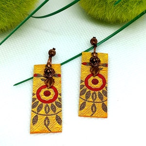 Peut inclure: Une paire de boucles d'oreilles pendantes jaunes avec un motif floral rouge et noir. Les boucles d'oreilles ont un charme de fleur de couleur cuivre et un crochet de couleur cuivre.