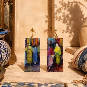 Può includere: Due orecchini rettangolari con un dipinto di persone in abiti colorati. Gli orecchini hanno ganci color oro. Lo sfondo include un vaso blu e bianco, una lanterna decorativa e una borsa fantasia.