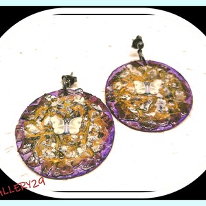 Op de afbeelding: Een paar paarse en gouden oorbellen met een wit vlinderontwerp. De oorbellen zijn gemaakt van metaal en hebben een getextureerd oppervlak.