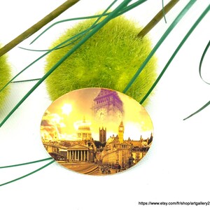 Peut inclure: Une épingle ronde en métal doré avec une image colorée de monuments londoniens, dont Big Ben et la cathédrale Saint-Paul. L'image est sur fond de ciel de coucher de soleil jaune et orange.