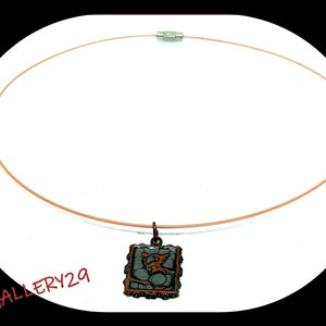 Puede incluir: Un collar con un colgante cuadrado que presenta un diseño abstracto gris y naranja. El colgante está enmarcado con un borde de metal negro y tiene un cierre plateado.