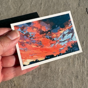 May include: A small, rectangular watercolor painting depicting a sunset. The artwork features vibrant hues of orange, red, blue, and dark navy, with a white border. The painting is held by a hand against a textured gray surface.
