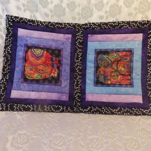 May include: A decorative pillow with a black and white patterned border. The pillow is divided into two sections, each featuring a colorful cat design with intricate details. The cat designs are framed with layers of purple, blue, and light purple fabric.