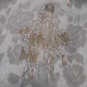 May include: A decorative tassel with a clear, beaded design. The tassel features a cluster of long, thin beads in shades of silver and gold, attached to a small, round, clear bead at the top. The tassel is likely used for embellishment.