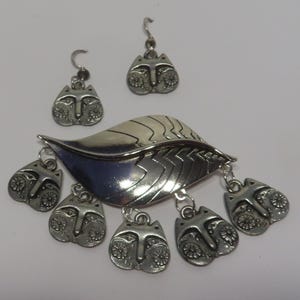 May include: Silver tone leaf-shaped pendant with five dangling charms, each featuring a stylized face. Two matching earrings with the same face design.