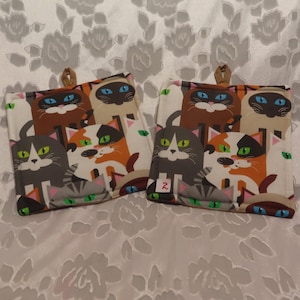 May include: Two cat-themed pot holders with a variety of colorful cats on a white background. The pot holders are square and have a loop for hanging. The text "2" is printed on one of the pot holders.