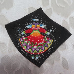 May include: A black fabric square with a colorful cat angel design. The cat is wearing a red, orange, yellow, green, purple, and blue dress with white stars and green leaves. The cat has a halo and wings. The background is black with white dots.