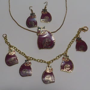 May include: A gold-tone jewelry set featuring a purple cat design. The set includes a necklace, a bracelet, and a pair of earrings.