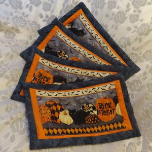 May include: Set of four Halloween-themed placemats. Each placemat features a dark blue border, an orange inner border, and a central design with pumpkins, black cats, and bats. One pumpkin has the text "Trick or Treat".