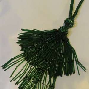 May include: A dark green beaded tassel with a loop for hanging. The tassel is made of long, thin beads, and the loop and top section are also beaded. The beads have a glossy finish.