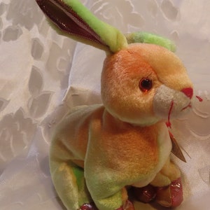 May include: A pastel green, orange, and pink tie-dye plush bunny with red eyes and a red nose. The bunny is sitting on a white surface.