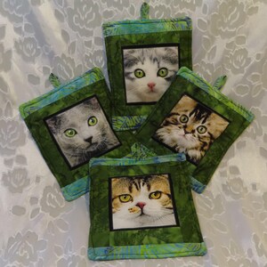 May include: Four green and blue fabric pot holders with different cat images. Each pot holder has a green background with a blue border and a white square in the center with a cat image.