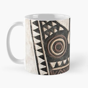 Puede incluir: Taza de cerámica blanca con un diseño geométrico en blanco y negro. El diseño presenta una cara estilizada con círculos concéntricos y triángulos.